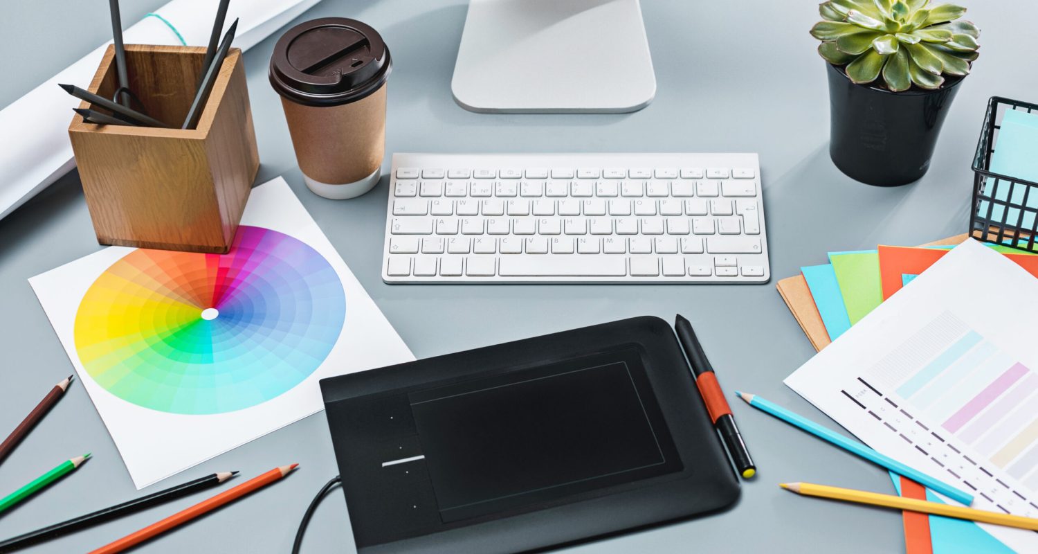 The desk with laptop, notepad with blank sheet, pot of flower, stylus and tablet for retouching. Workspace of professional retoucher. Creativity, design, art and modern equipments concept